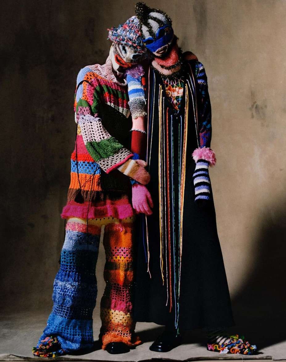 Two people in colorful knitwear and masks pose together in a studio setting against a neutral background, showcasing high fashion and artistry.