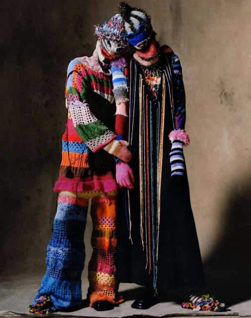 Two people in colorful knitwear and masks pose together in a studio setting against a neutral background, showcasing high fashion and artistry.