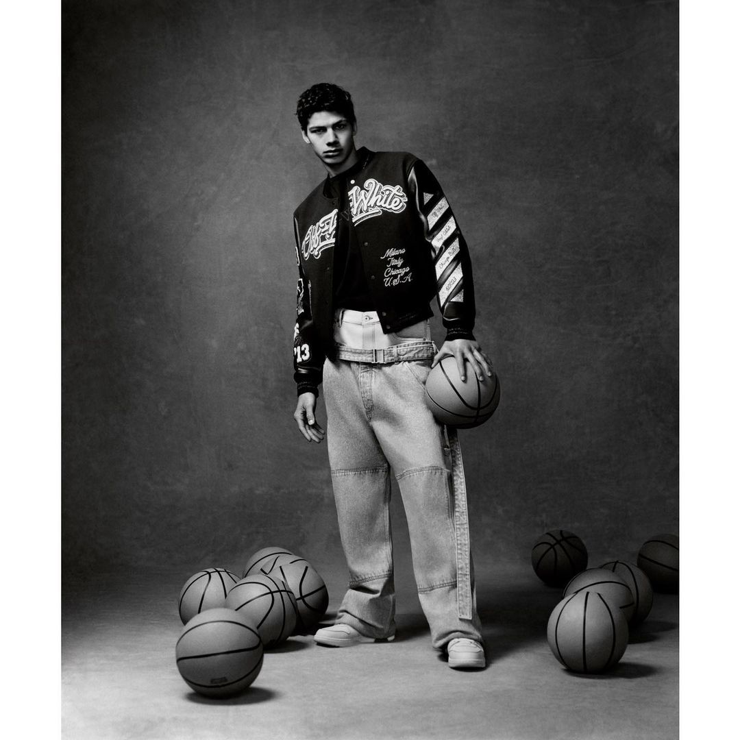 Portrait of a young adult man posing with a basketball wearing a black and white jacket and jeans, with basketballs around him on grey background.