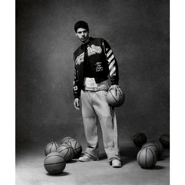 Portrait of a young adult man posing with a basketball wearing a black and white jacket and jeans, with basketballs around him on grey background.