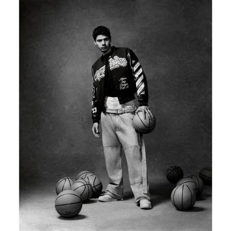 Portrait of a young adult man posing with a basketball wearing a black and white jacket and jeans, with basketballs around him on grey background.