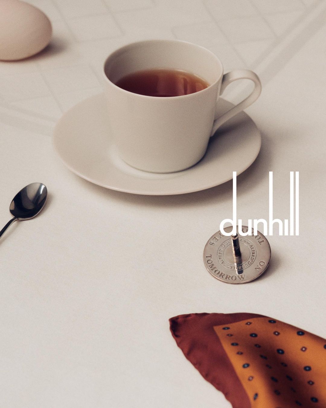 Overhead shot of a white ceramic cup with tea, a spoon, scarf, a coin, and the Dunhill logo on a white surface.