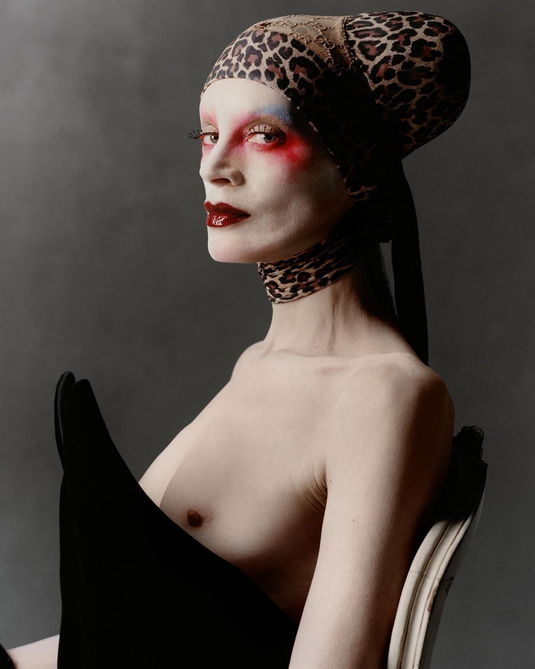 Striking fashion portrait: bare-chested adult female with painted face, leopard-print head wrap, and dramatic dark makeup against a grey backdrop.