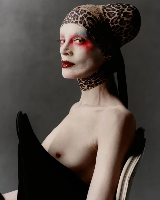 Striking fashion portrait: bare-chested adult female with painted face, leopard-print head wrap, and dramatic dark makeup against a grey backdrop.