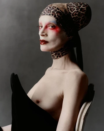 Striking fashion portrait: bare-chested adult female with painted face, leopard-print head wrap, and dramatic dark makeup against a grey backdrop.