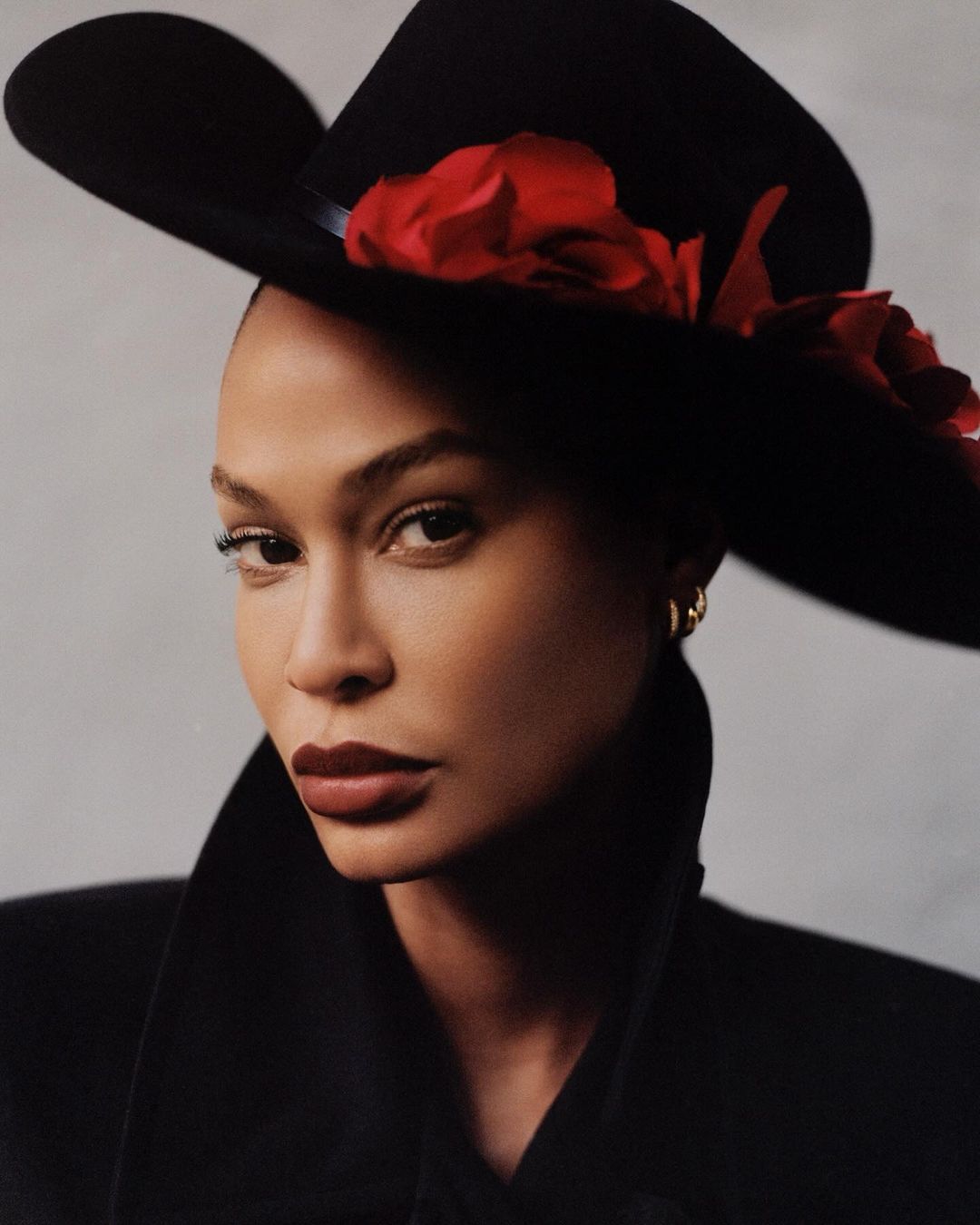 Close-up portrait of an adult female wearing a black hat with red floral accents and a black jacket against a neutral background.