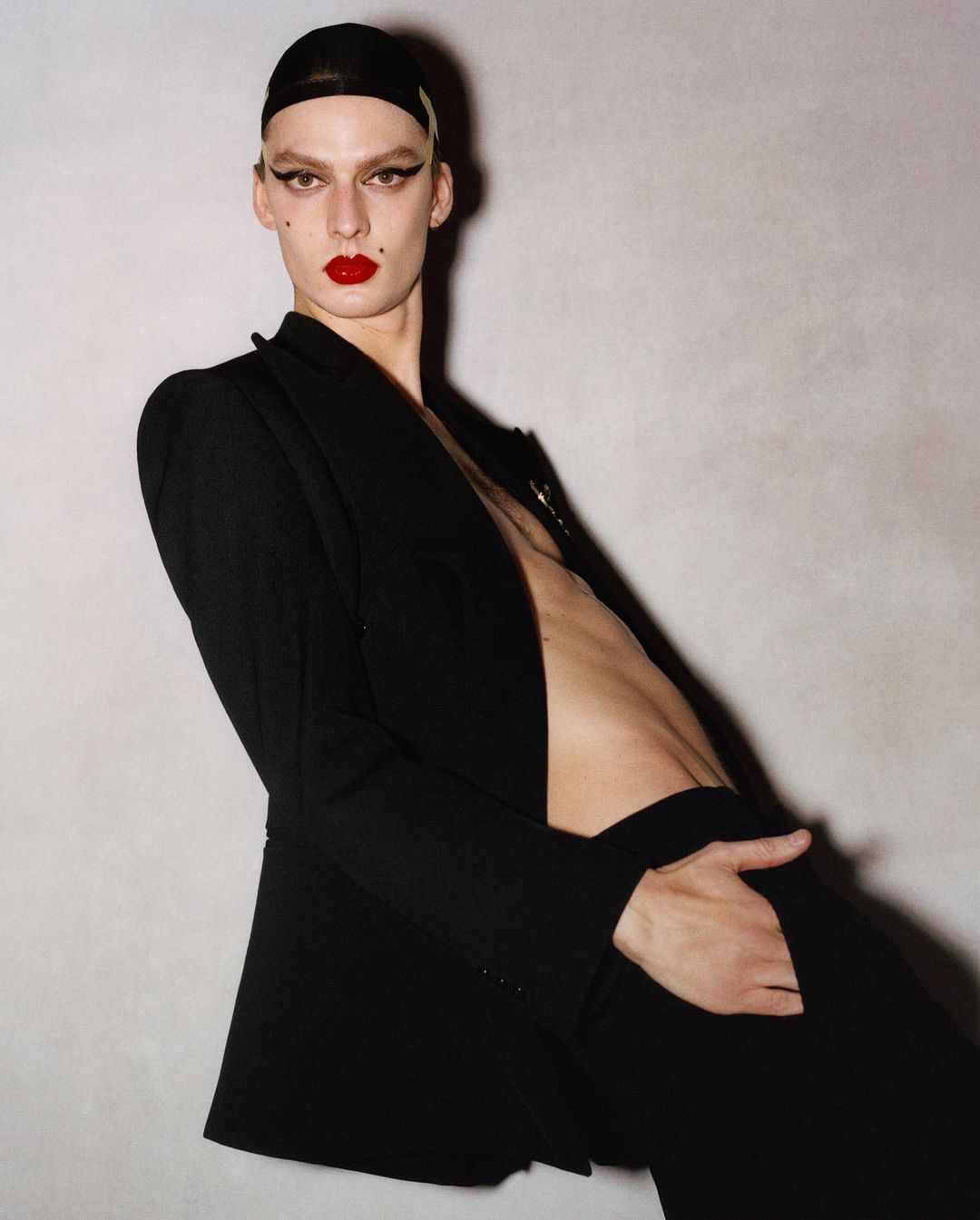 Person in black business suit and black cap poses against a neutral wall, featuring red lipstick and prominent cheekbones.