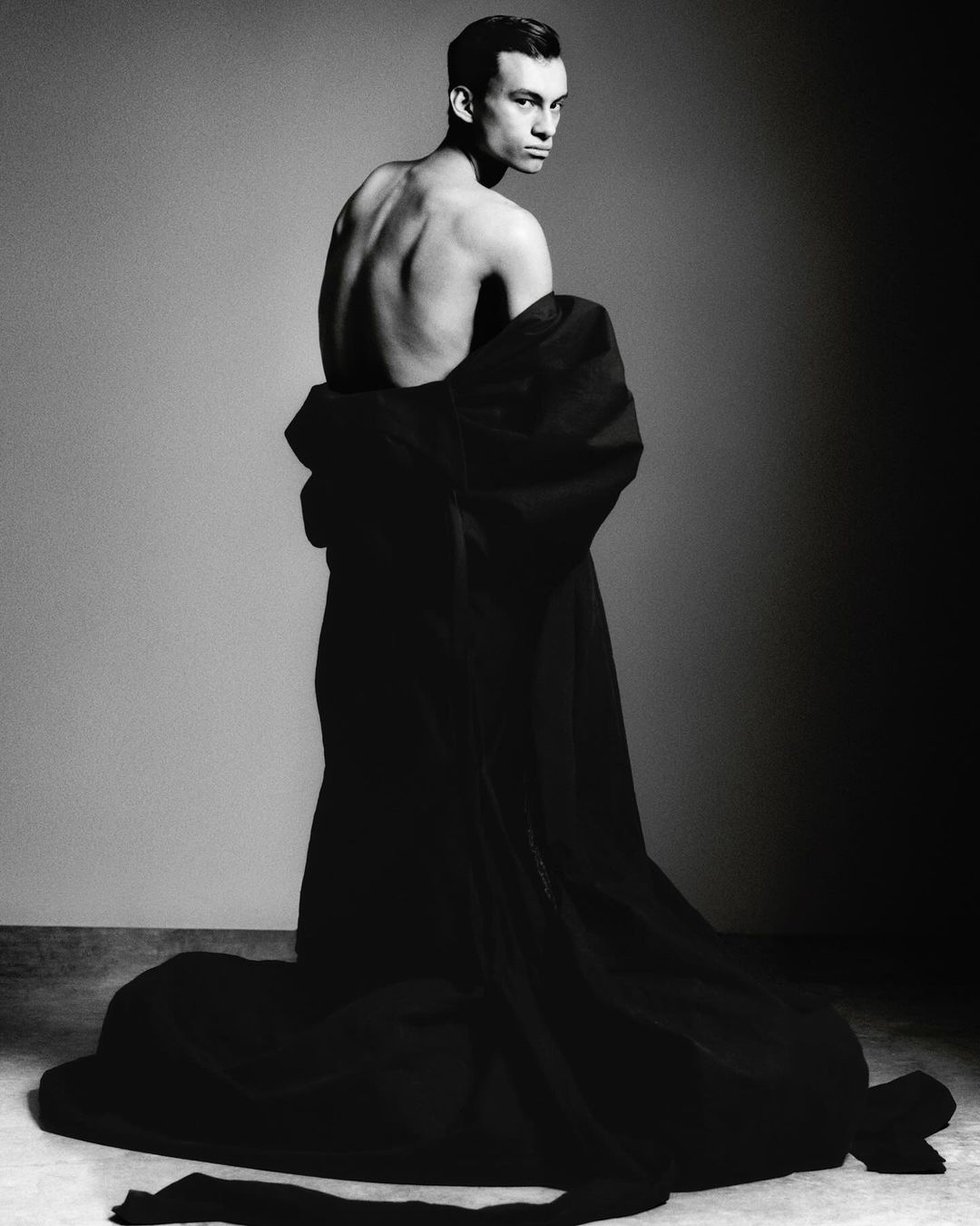 Monochrome portrait of a slender, shirtless male model draped in dark fabric looking over his shoulder in a studio setting.