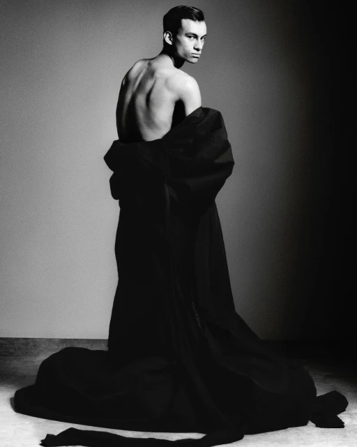 Monochrome portrait of a slender, shirtless male model draped in dark fabric looking over his shoulder in a studio setting.