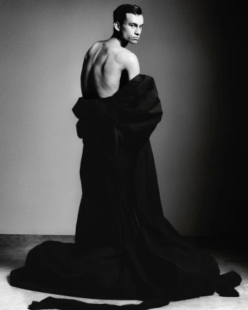 Monochrome portrait of a slender, shirtless male model draped in dark fabric looking over his shoulder in a studio setting.