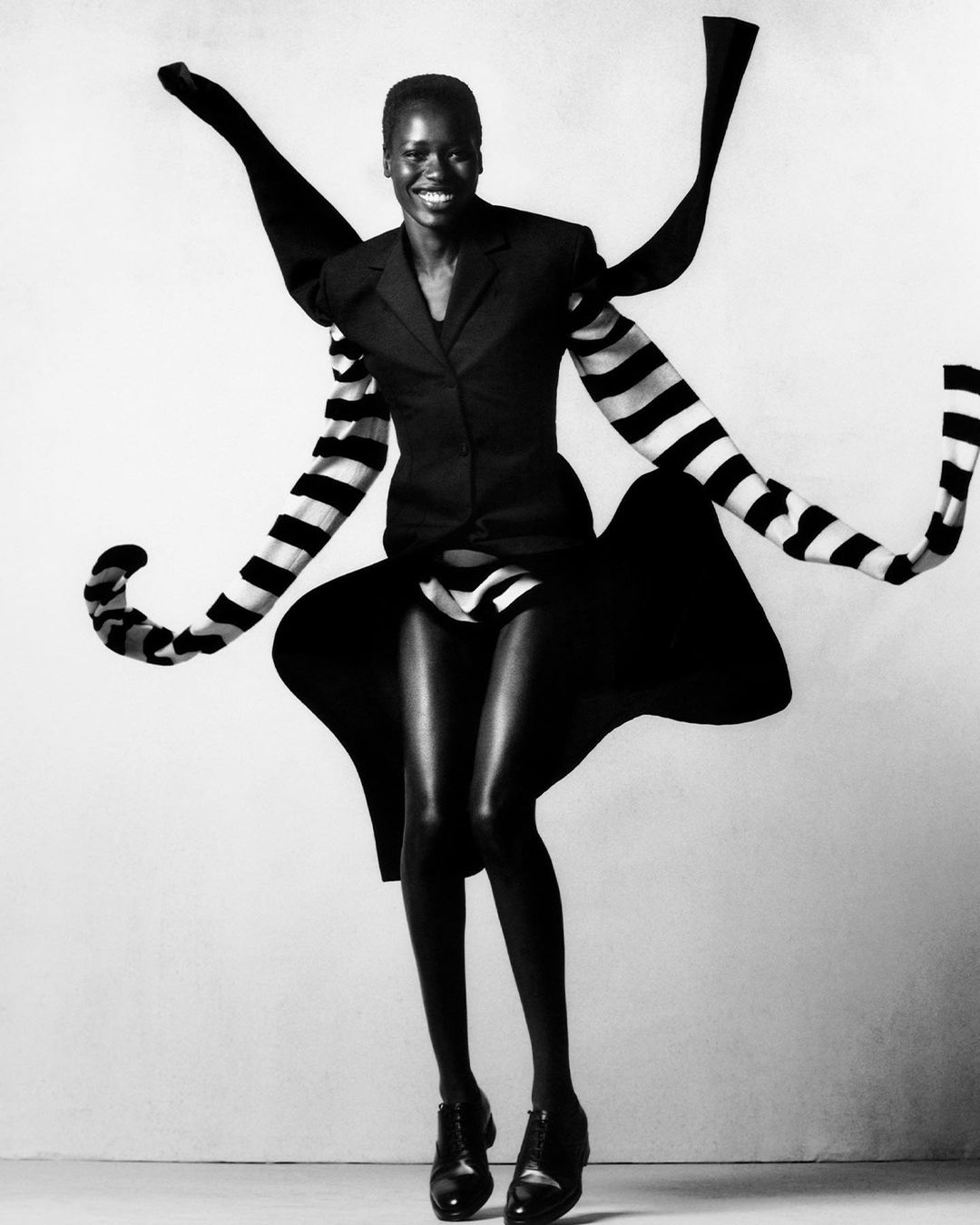 Elegant black model in a blazer, striped sleeves, and dress strikes a pose against a white background in a studio fashion shoot.
