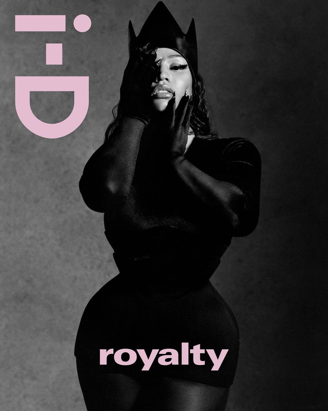 Black and white portrait of a woman with her hands on her face, wearing a black crown, gloves, and dress against a grey backdrop.