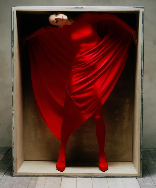 A woman in a red dress and heels poses inside of a wood box. Her red dress is draped around her arms and legs, contrasting with the box.