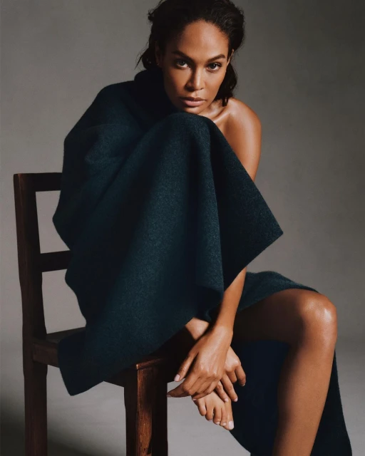 Portrait of a dark-skinned woman with shoulders and bare legs sitting in a dark green garment on a wood chair in a gray studio.