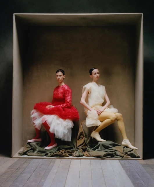 Two women in stylized sheer dresses sit inside a square box on a set with a painted backdrop and wooden floor. Fashion shoot.