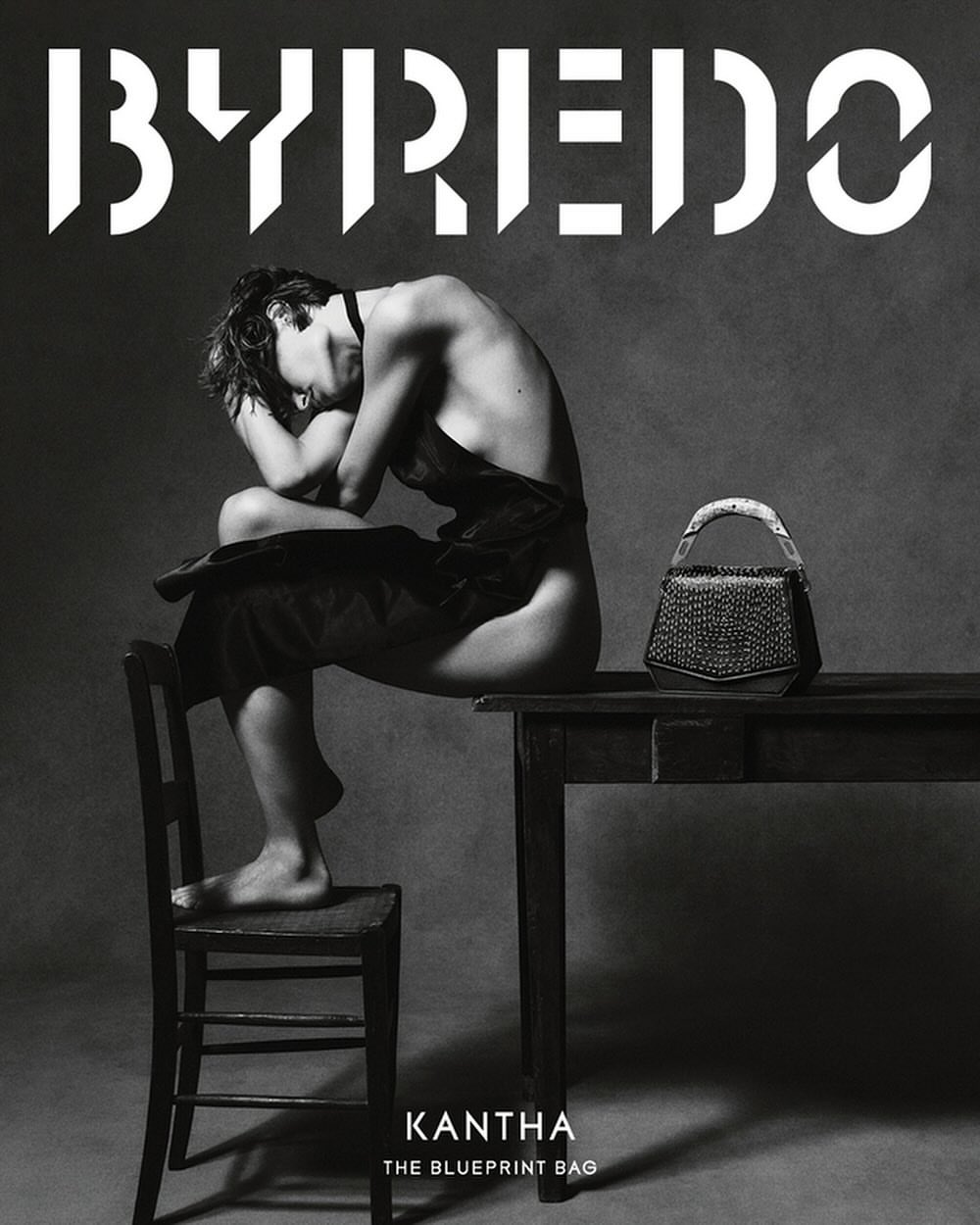 Black and white studio shot of a partially nude adult sitting on a chair next to a table with a dark-colored handbag, with branding in the background.