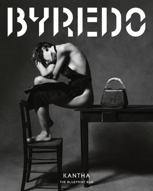 Black and white studio shot of a partially nude adult sitting on a chair next to a table with a dark-colored handbag, with branding in the background.