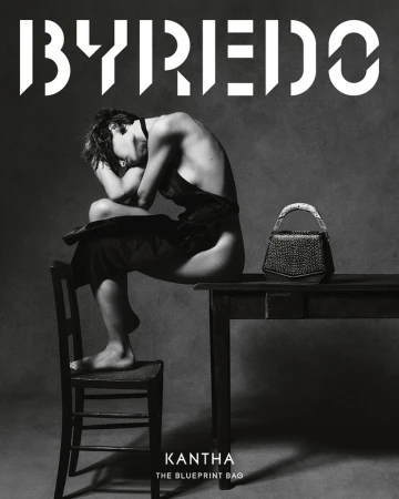 Black and white studio shot of a partially nude adult sitting on a chair next to a table with a dark-colored handbag, with branding in the background.
