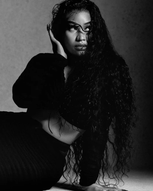Monochrome portrait of an adult female model with long curly hair wearing black crop top and skirt, posing against a textured gray background.