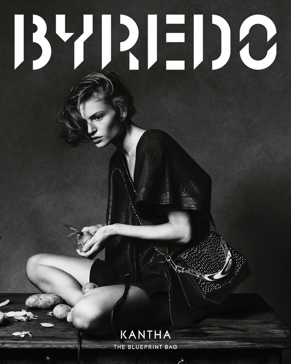 Black and white fashion shot: a model in a dark dress sits cross-legged, peeling potatoes, showcasing a studded handbag on a rustic surface.