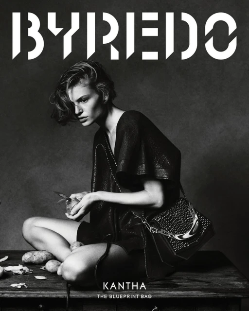 Black and white fashion shot: a model in a dark dress sits cross-legged, peeling potatoes, showcasing a studded handbag on a rustic surface.