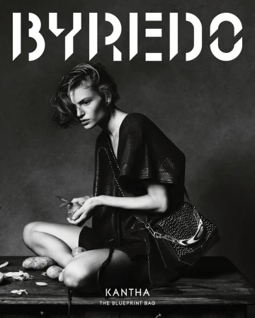 Black and white fashion shot: a model in a dark dress sits cross-legged, peeling potatoes, showcasing a studded handbag on a rustic surface.