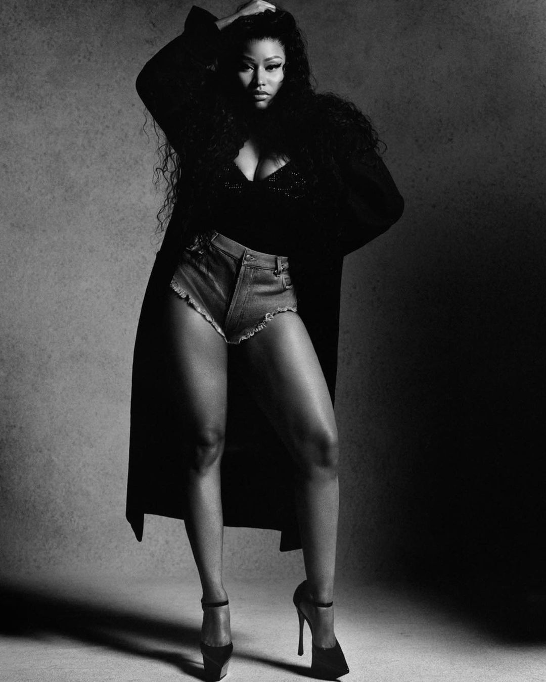 Monochrome portrait of Nicki Minaj wearing a dark coat, denim shorts, and high heels against a plain textured backdrop.