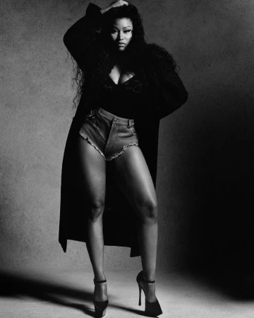 Monochrome portrait of Nicki Minaj wearing a dark coat, denim shorts, and high heels against a plain textured backdrop.
