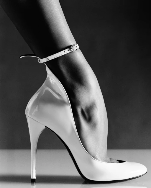 Close-up studio shot of a woman's foot in a white patent leather stiletto heel with an ankle strap, set against a dark grey background.