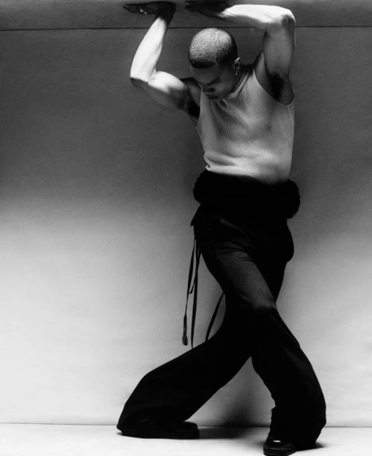 Monochrome studio shot of a muscular adult male in tank top and draped trousers, pressing upwards with hands against an unseen surface.