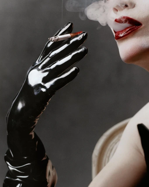 Woman with red lips wearing a black glove holds a lit cigarette with smoke against a grey background in a beauty shot.