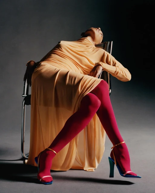 Fashion photo of a bald woman in a long beige dress sitting in a metal chair with red tights and blue heeled sandals in a studio.