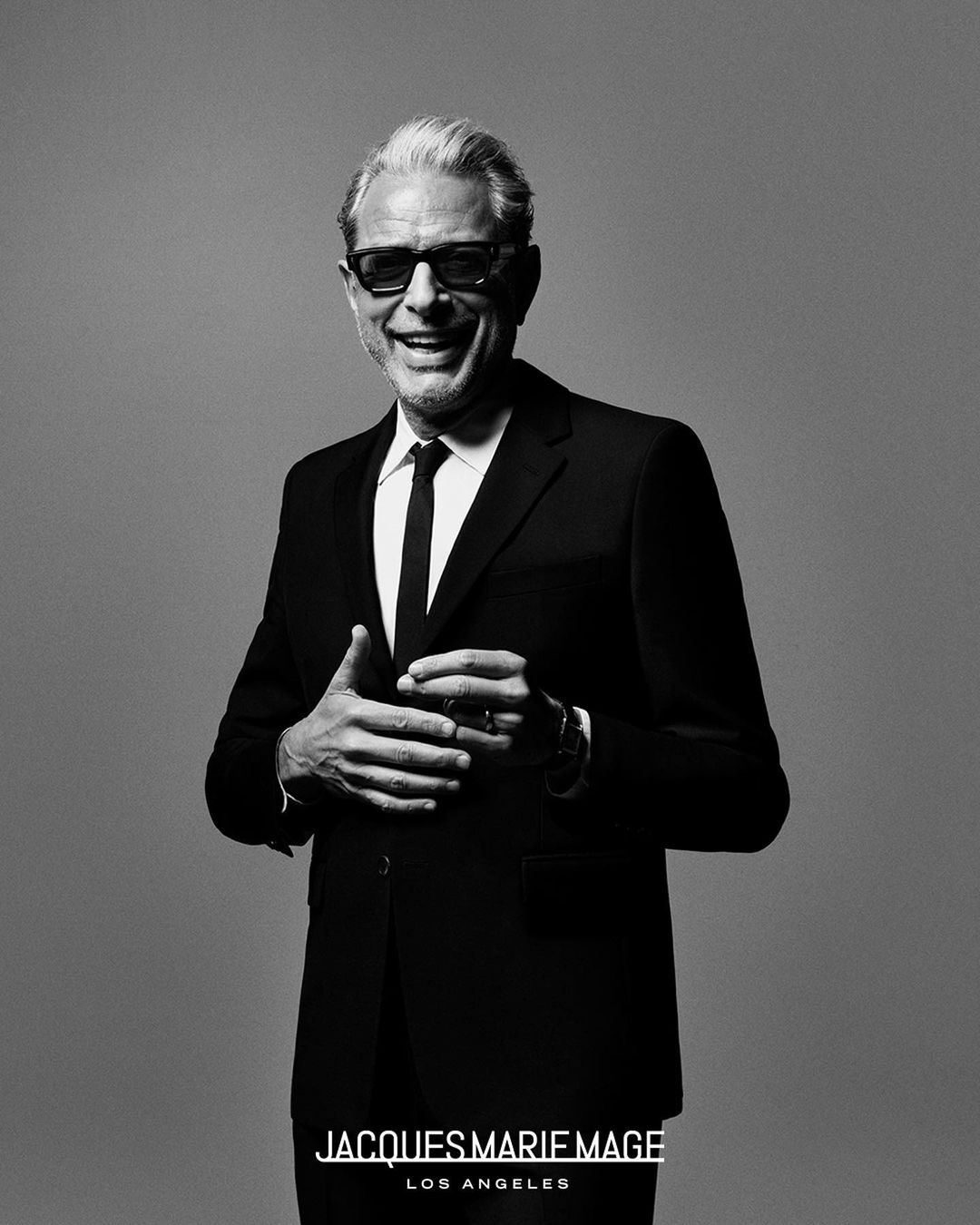Jeff Goldblum in a dark suit and glasses smiling against a grey background by Luis Alberto Rodriguez