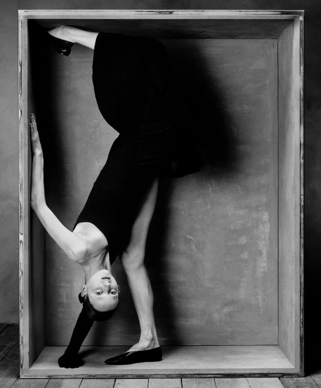 Black-and-white photo of a woman in a black dress contorting her body inside of a wooden box, creating a visually striking and unique pose.