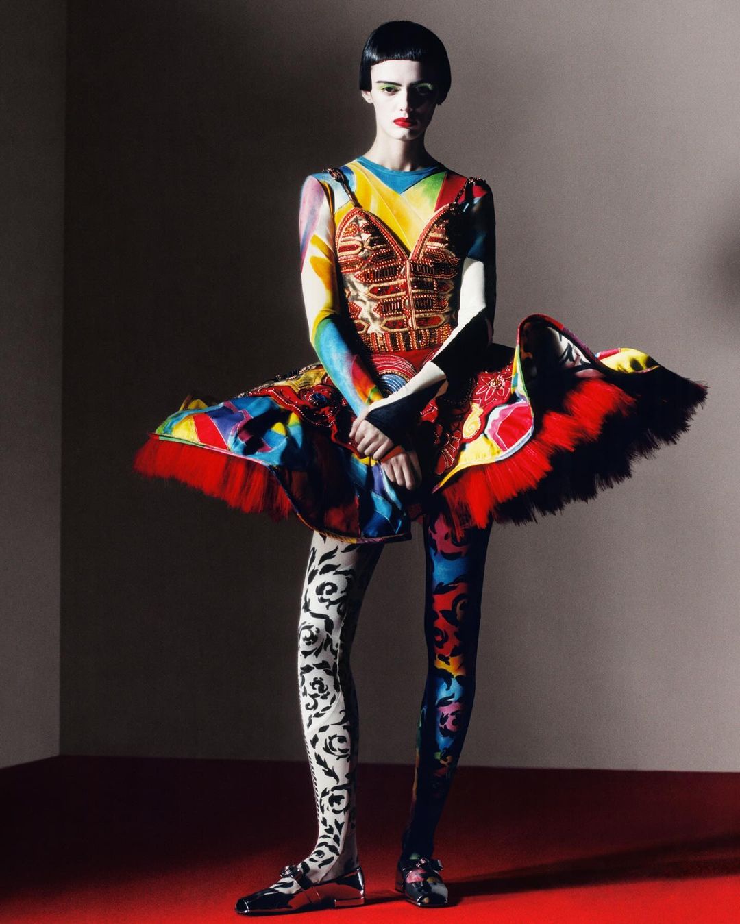 Fashion model poses in colourful, patterned dress with a dramatic tutu-style skirt, leggings, and black shoes against a studio backdrop.