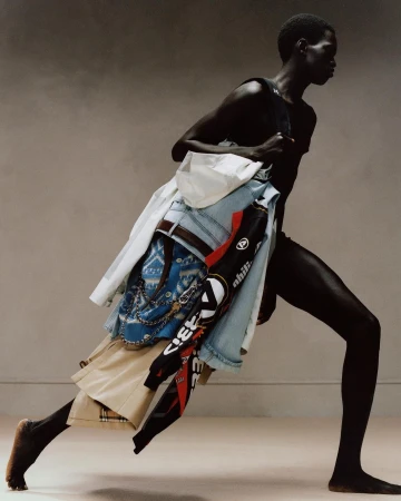 Model carrying a layered pile of garments including denim jeans, white shirt, and patterned jacket.