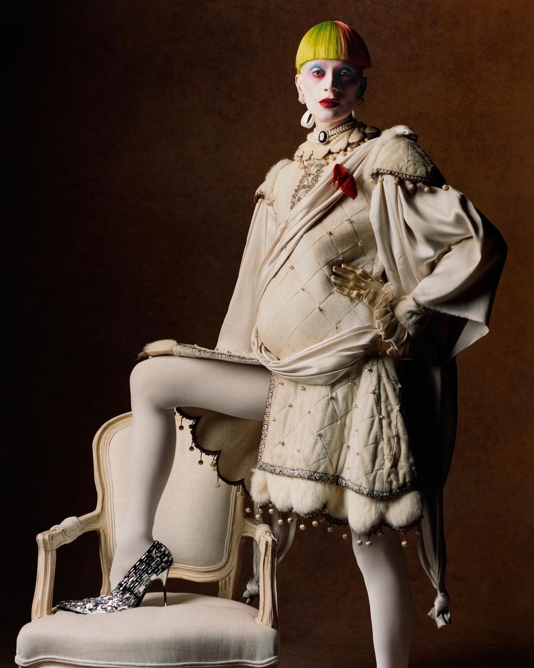 A fashion model posing in a white renaissance-style garment with pearl accents while leaning on an antique chair, set against a brown backdrop.