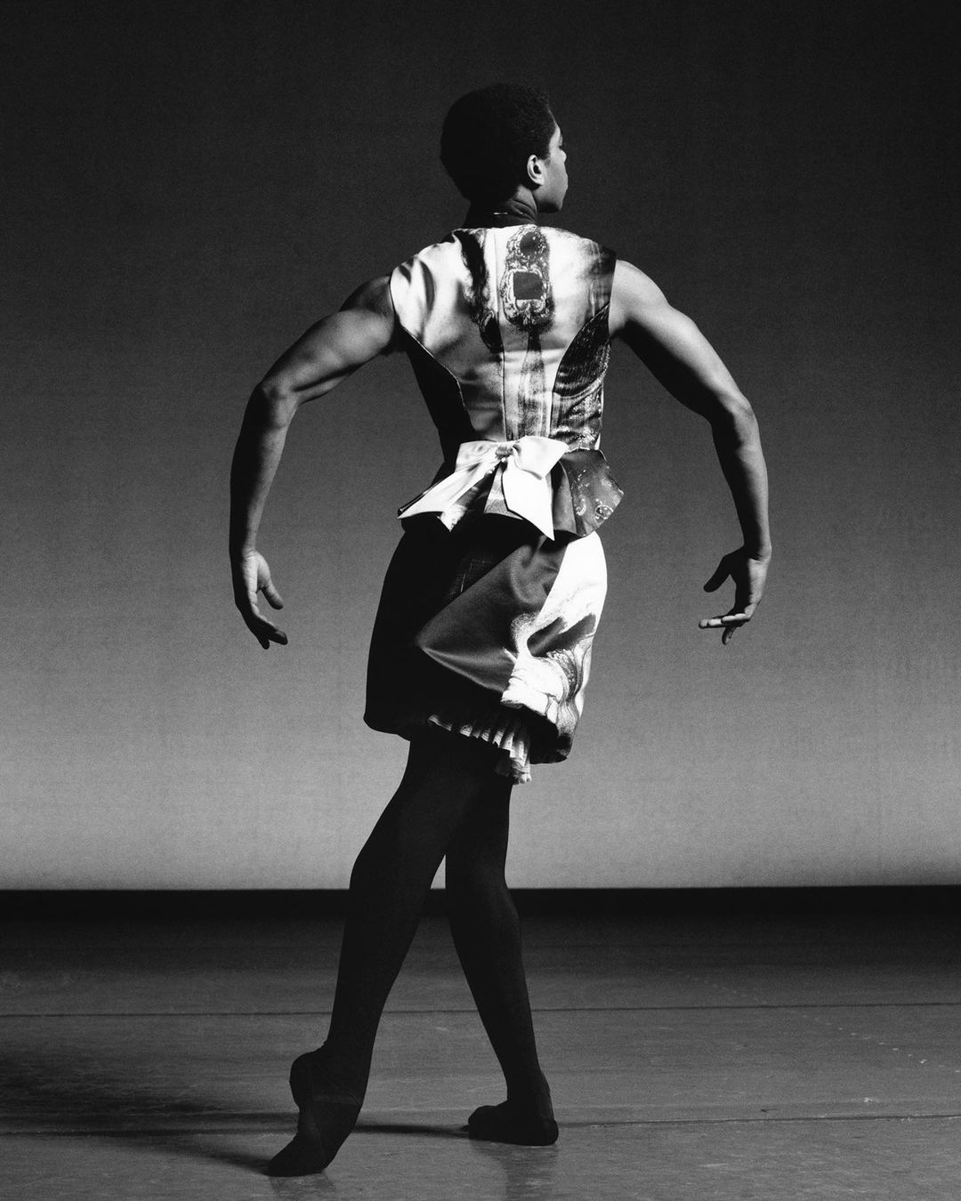 An African-American ballet dancer poses in black and white wearing a decorative sleeveless top and tights on a grey stage background.