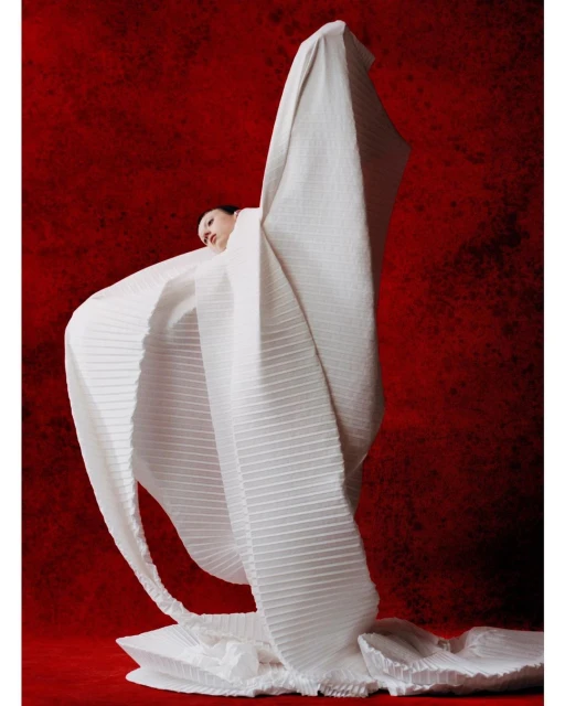 Elegant woman in draped white fabric against a red textured background, showcasing fashion and artistic photography.