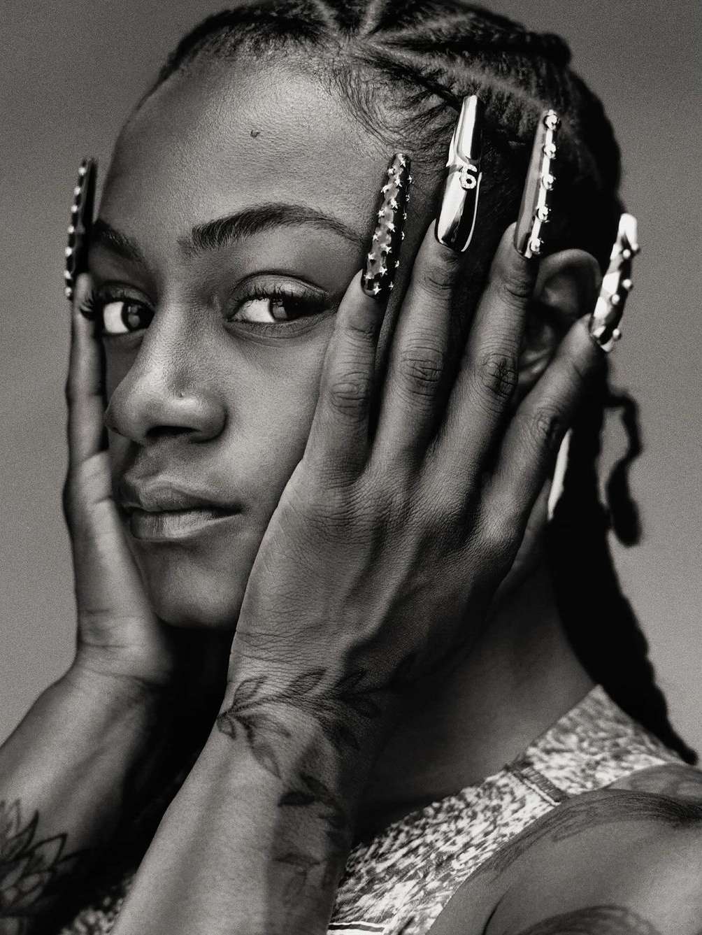 Close-up portrait of a young woman with hands framing her face, showcasing braided hair, long decorative nails, and a patterned top.