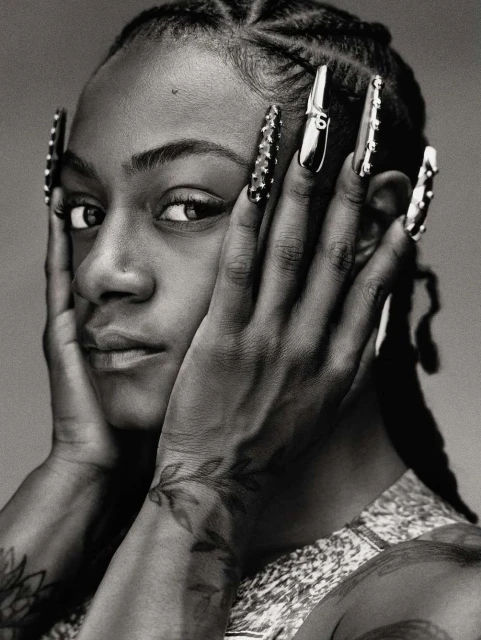 Close-up portrait of a young woman with hands framing her face, showcasing braided hair, long decorative nails, and a patterned top.