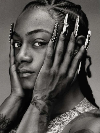 Close-up portrait of a young woman with hands framing her face, showcasing braided hair, long decorative nails, and a patterned top.