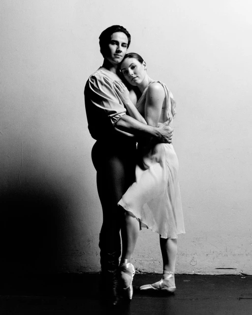 A pair of ballet dancers in costume embrace closely in a studio setting, set against a stark white wall in soft, monochrome light.