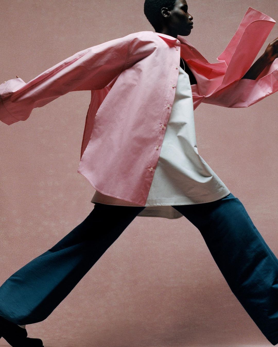 Fashion photo of a Black woman walking in a pink button-down shirt, white top, and blue pants against a pink background in studio lighting.