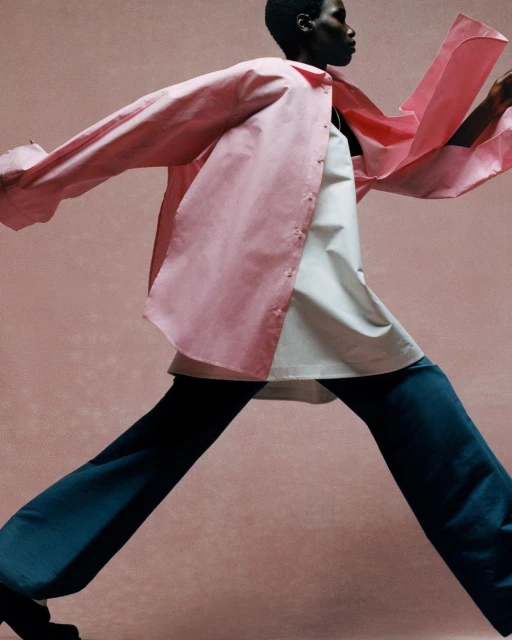 Fashion photo of a Black woman walking in a pink button-down shirt, white top, and blue pants against a pink background in studio lighting.