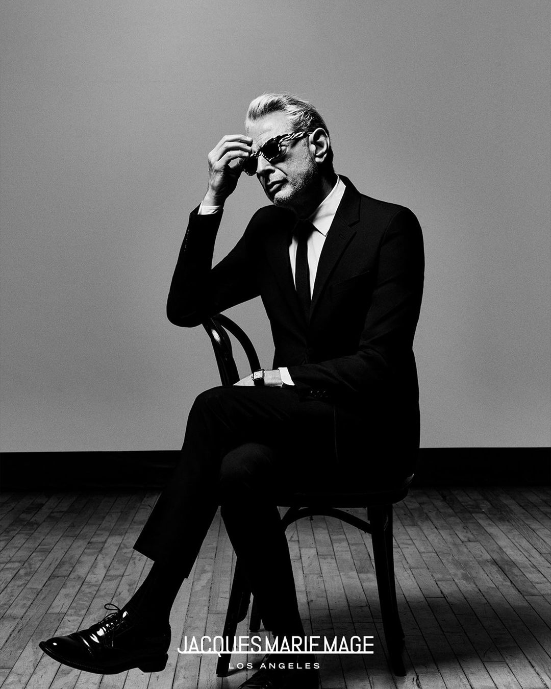 Black-and-white portrait of an older male model wearing sunglasses, a suit, and tie, seated on a chair in a photo studio.