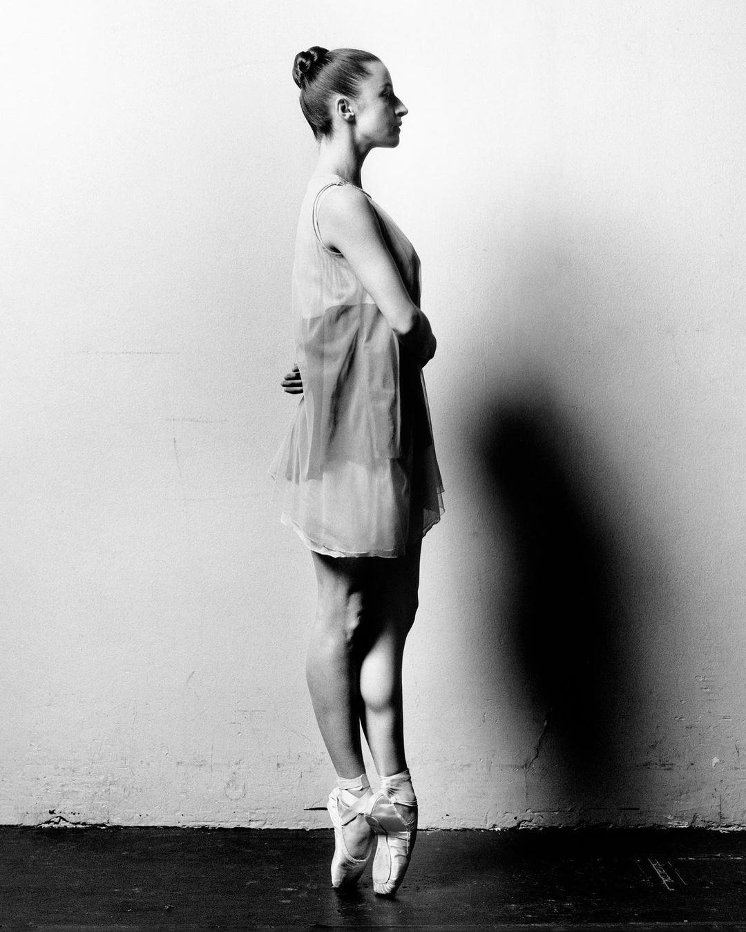 Ballet dancer en pointe in a studio. Side view shows a young woman with her arms behind her back, wearing a dress and ballet shoes.