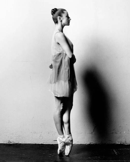 Ballet dancer en pointe in a studio. Side view shows a young woman with her arms behind her back, wearing a dress and ballet shoes.
