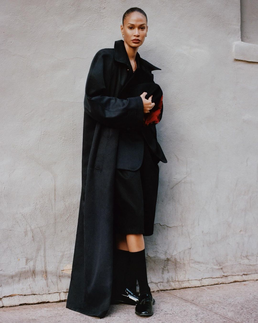 Model wearing an oversized black coat and dark loafers posing against a textured grey wall.