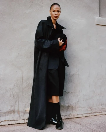 Model wearing an oversized black coat and dark loafers posing against a textured grey wall.